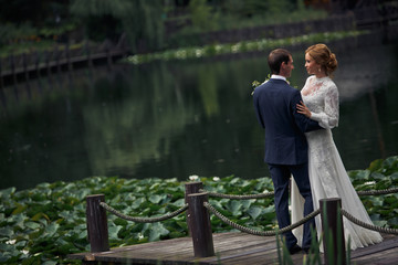 Groom holds delicate bride's waist standing with her on a bridge