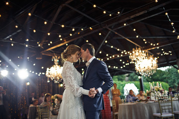 Lamps sparkle behind a wedding couple dancing at the first time