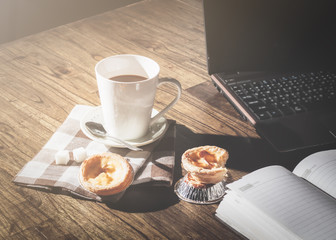 Egg tart, coffee, notebook, and laptop on wooden table in morning sunlight.