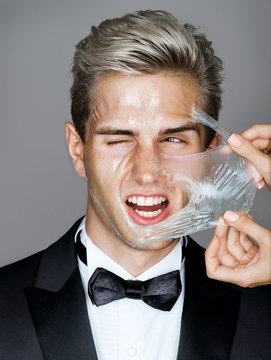 Pain. Elegant man in black suit removes peeling off a facial mask. Photo disaffected rich man on gray background. Skin care concept