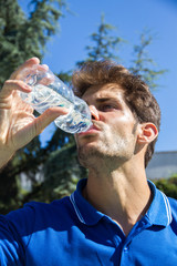 Professional tennis player wearing a blue polo is resting tennis match drinking water from a bottle. He is sitting in front of the tennis courts. 