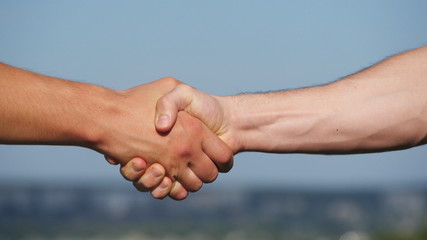 Friendly handshake of two unrecognizable muscular white men on blue sky background. Shaking of male arms outdoor. Two strong men having firm handshake outside. Teamwork and friendship. Close-up