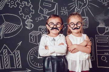 Two little girls as with mustache and glasses standing on dark background of painted wall. Their hands folded