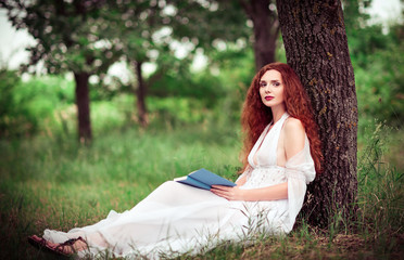 Lovely redhead woman sitting under tree and reading a book