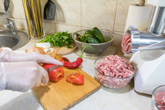 Woman is cutting pepper on a wooden cutting board in the modern kitchen surrounded by fresh products, fresh vegetables and pork meat in a glass bowl.