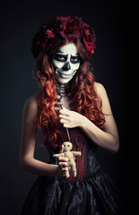Young voodoo witch with muertos makeup (sugar skull) holds voodoo doll and needle