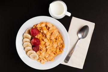Corn flakes with slice banana and strawberry in bowl, milk in jar, spoon on napkin.