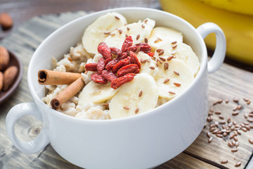 Oatmeal porridge with super foods: dry goji berries, flax seeds, cinnamon, banana and almonds in white bowl. Close up, angle view