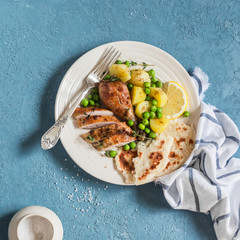 Lemon thyme baked chicken, potatoes and green peas on a white plate on a blue background. Delicious lunch