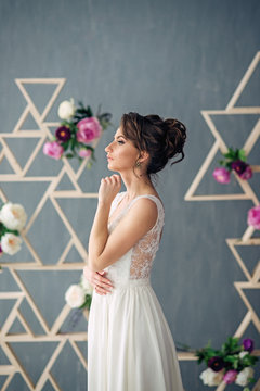 Beautiful young bride in a wedding dress looks out the window with gray wall and pink flowers on the background