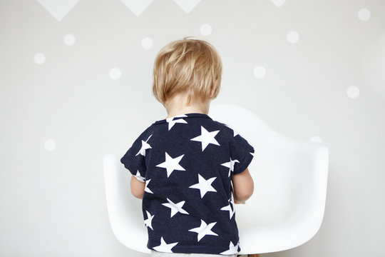 Back view of little Caucasian boy with blonde hair wearing t-shirt with stars on it, playing with toys in nursery. Cute toddler standing in front of white chair, spending day at home. Visual effects
