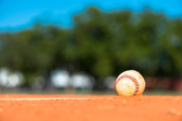 Worn little league baseball on red dirt pitchers mound with outfield fence and trees in background