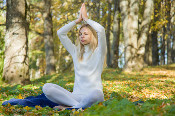 In autumn day young woman practicing yoga in the park atmosphere, where sunlight plays with shadows. Fallen yellow leaves. White clothes.