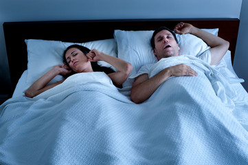 Snoring man sleeping snoring from obstructive sleep apnea in bed at night with tortured wife plugging ears with fingers