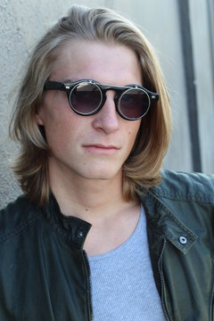 Handsome young man with blond long hair and sunglasses isolated on grey background. Fashion studio shot.