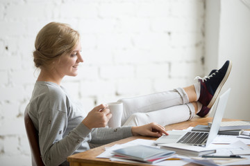 Profile portrait of a smiling student girl working with a laptop, her legs on the desk, mug in her hand. Education concept photo, lifestyle