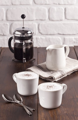 Cappuccino cups, french press and creamer are on the wooden table.