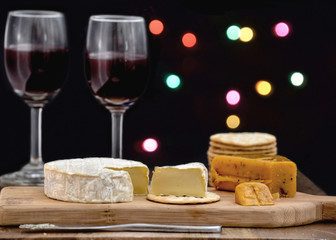Brie and hot pepper jack cheese on wooden cheese board, accompanied by crackers and two glasses of red wine, against black background decorated with colorful holiday lights