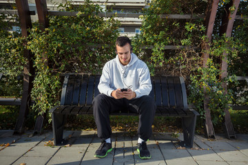 Attractive young man is relaxing after workout in a park. He is sitting on the bench and using mobile phone for listening to music on headphones.