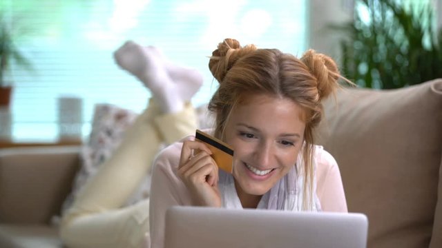 A young woman uses a golden credit card online lying on the couch.