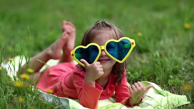 Funny little girl in big sunglasses in the shape of hearts lies on green grass. Funny girl show his tongue. Beautiful little girl in glasses lies on the green grass and looks at the camera