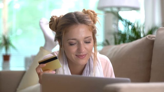 A young woman uses a golden credit card online lying on the couch.