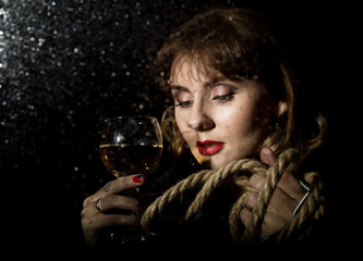 mysterious young woman with a glass of wine posing behind transparent glass covered by water drops. on a dark background