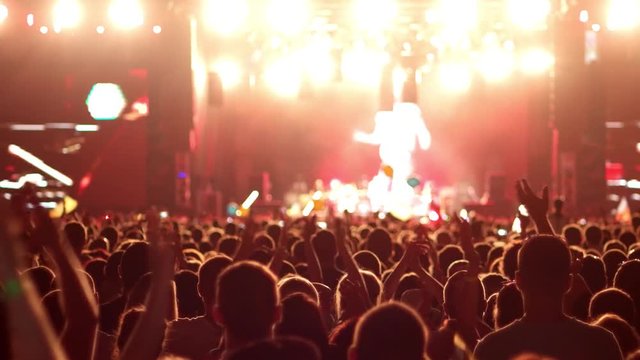 A crowd of people at a concert, a show of hands waving, clap rock musicians on the brightly lit stage, on the big screens in the background can be seen artists, bright multi colored lights on stage
