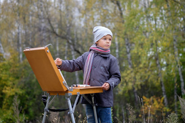 Юный живописец  с мольбертом в осеннем лесу пишет картину 