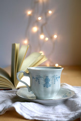 Tea in a vintage porcelain cup and open book. Fairy lights in the background. Selective focus.