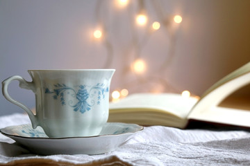 Tea in a vintage porcelain cup and open book. Fairy lights in the background. Selective focus.