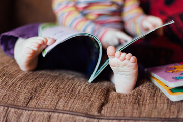 Toddler girl sitting on brown sofa reading a book with focus on her feet