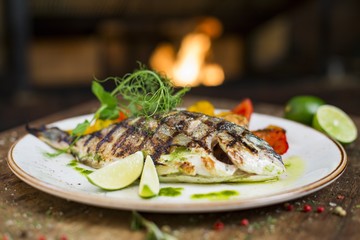 Main course gourmet fish - delicious grilled golden sea bream or dorade with vegetable garnish served on a wooden table, fireplace on background