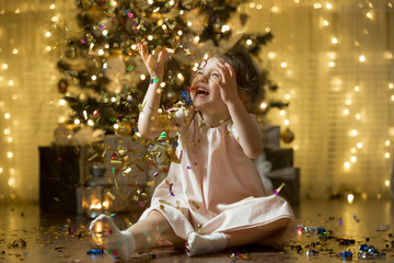 Excited little girl laughing, Catching confetti. Beautifully decorated christmas tree and house with lights and lantern, christmas gifts. 