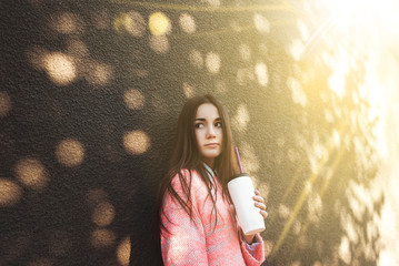 Cute young girl in pink sweater holding a cup with a straw. Sunny day. Funny mood.