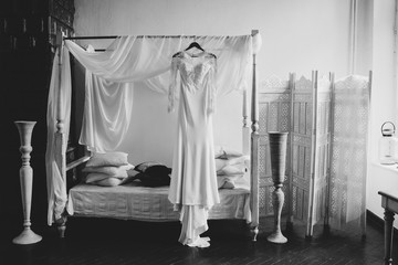 black and white image of a lace and silk wedding dress hang on vintage bed during the bride's morning preparation