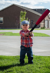 Toddler boy playing baseball with a white wiffle ball and red bat. Wearing blue motoring cap, red plaid shirt and blue jeans on grass on sunny cloudy day.