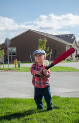Toddler boy playing baseball with a white wiffle ball and red bat. Wearing blue motoring cap, red plaid shirt and blue jeans on grass on sunny cloudy day.