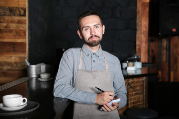 Bartender with notebook and pen standing near wooden bar counter