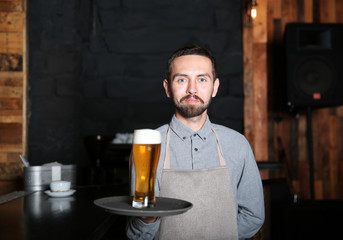 Bartender holding tray with glass of beer near wooden bar counter