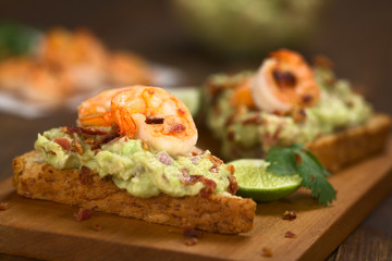 Vollkorntoastbrot belegt mit Guacamole (Avokado-Creme), gebratenen Garnelen und Speckstückchen (Selektiver Fokus, Fokus auf die Vorderseite der Garnele auf dem Brot)