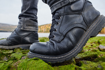 Legs in leather boots stand on the banks of the river of stones, moss-covered, mountains in the background with trees closeup