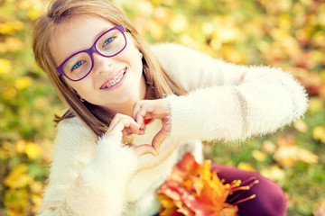 Smiling little girl with braces and glasses showing heart with hands.Autum time.