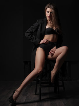 Seductive, beautiful and attractive young woman with loose brown hair, sexy gorgeous figure and long legs in the black seamless underwear and wool coat is posing on the chair in the studio