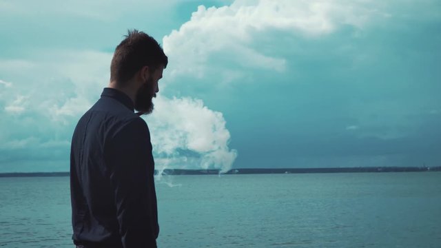 Young stylish man with a beard smokes an electronic cigarette in the background of the sea and clouds epic. Modern fashion vaping