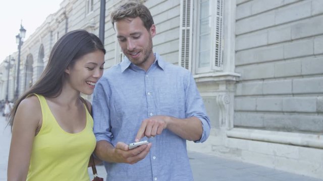 Couple using mobile cell smart phone in city outdoors. Man showing smartphone screen laughing happy outside. Asian woman, Caucasian man. RED EPIC SLOW MOTION.