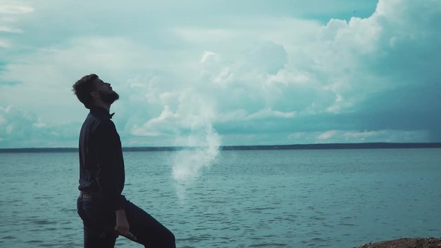 Young stylish man with a beard smokes an electronic cigarette in the background of the sea and clouds epic. Modern fashion vaping