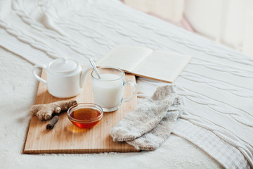 Obraz na płótnie Canvas Hot milk in a glass cup and honey on a wooden board. Treatment of children a hot drink. Treatment of folk remedies in bed. Warm knitted socks. The book to read.