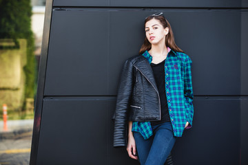 Portrait of brunette girl wearing leather jacket standing outdoors in the city against the black urban wall