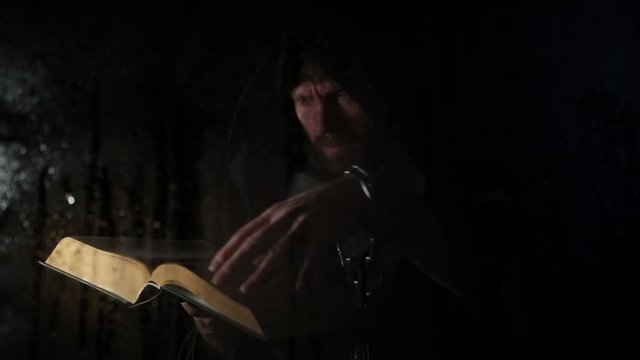 necromancer casts spells from thick ancient book by candlelight, behind transparent glass covered by water drops on a dark background